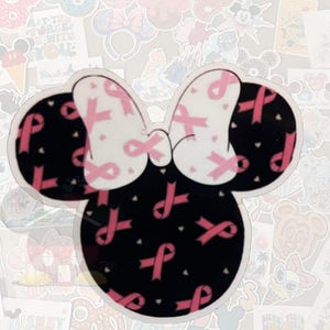 K&ouml;nnte beinhalten: Ein Minnie-Mouse-f&ouml;rmiger Aufkleber mit schwarzem Kopf und Ohren und einer wei&szlig;en Schleife. Die Schleife ist mit rosa B&auml;ndern verziert, und der Kopf ist mit rosa B&auml;ndern und kleinen wei&szlig;en Herzen bedeckt. Der Aufkleber ist von anderen Aufklebern umgeben.