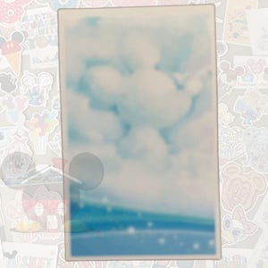 K&ouml;nnte beinhalten: Ein rechteckiges, helles Objekt mit einem verschwommenen Bild eines blauen Himmels mit wei&szlig;en Wolken. Der untere Teil des Bildes hat einen blauen und gr&uuml;nen Farbverlauf. Das Objekt ist von verschiedenen bunten Aufklebern umgeben.