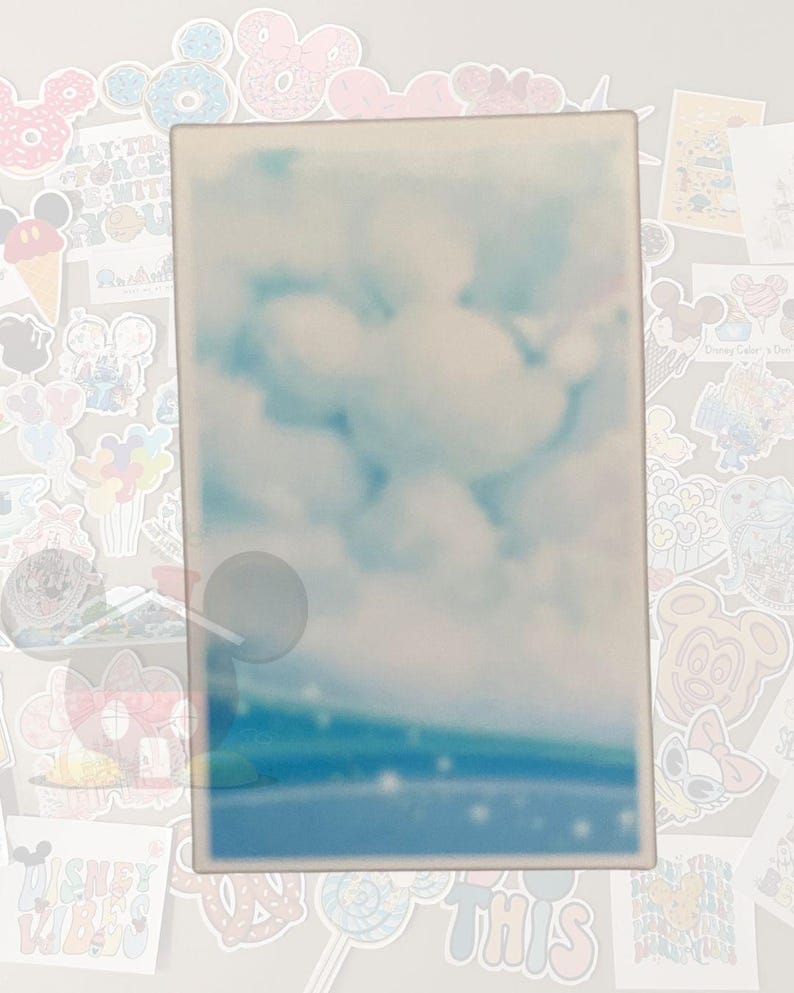 K&ouml;nnte beinhalten: Ein rechteckiges, helles Objekt mit einem verschwommenen Bild eines blauen Himmels mit wei&szlig;en Wolken. Der untere Teil des Bildes hat einen blauen und gr&uuml;nen Farbverlauf. Das Objekt ist von verschiedenen bunten Aufklebern umgeben.