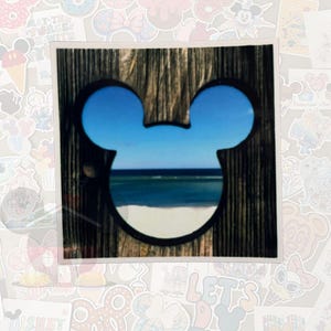 May include: A square sticker featuring a wooden frame with a Mickey Mouse head-shaped cutout. The cutout reveals a beach scene with blue sky, ocean, and white sand. The sticker is surrounded by other colorful stickers.