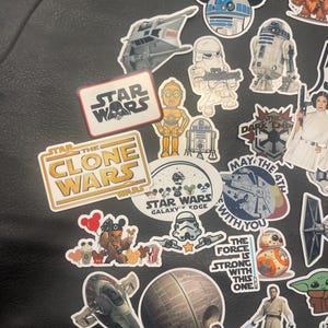May include: A collection of Star Wars themed stickers featuring characters like R2-D2, C-3PO, and Yoda. The stickers include various designs, including the Star Wars logo, the Death Star, and the Millennium Falcon. The stickers are on a black surface.