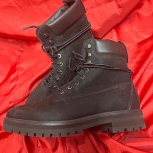 Puede incluir: Un par de botas de trabajo de cuero marrón oscuro con cordones. Las botas tienen un cuello acolchado, una suela con dibujo y el logotipo de Timberland. Están sobre un fondo rojo.
