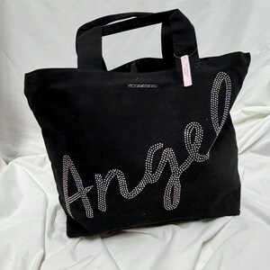 May include: Black tote bag with the word "Angel" spelled out in sparkling rhinestones. The bag has two handles and the words "Victoria's Secret" are printed on the top. A pink tag hangs from one of the handles.