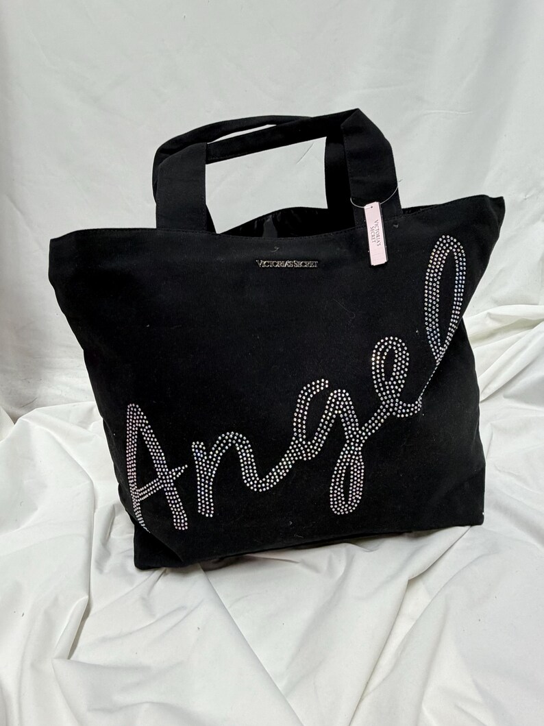 May include: Black tote bag with the word "Angel" spelled out in sparkling rhinestones. The bag has two handles and the words "Victoria's Secret" are printed on the top. A pink tag hangs from one of the handles.