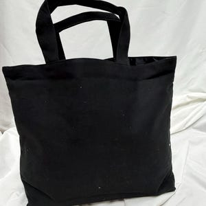 May include: A black tote bag with two sturdy handles. The bag is made of a solid black fabric and has a rectangular shape. The handles are attached to the top of the bag, allowing it to be carried by hand or over the shoulder.