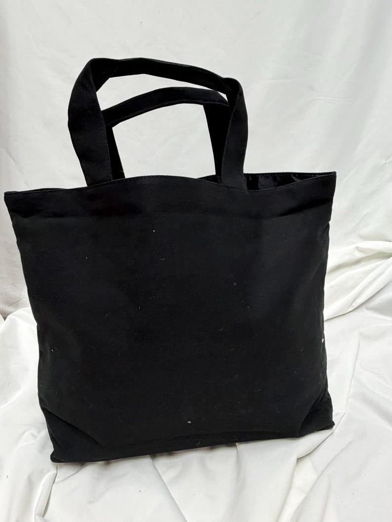 May include: A black tote bag with two sturdy handles. The bag is made of a solid black fabric and has a rectangular shape. The handles are attached to the top of the bag, allowing it to be carried by hand or over the shoulder.