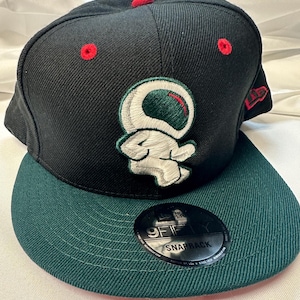 May include: Black and green snapback cap with a flat peak. The cap has a white and green embroidered design, red stitching on the crown, and a New Era logo. The peak is dark green.