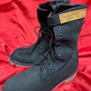 Puede incluir: Botas Timberland negras con cordones y una etiqueta de cuero color canela. Las botas tienen una suela resistente y un puño plegable. La etiqueta dice "Timberland 03885-NH".