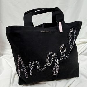 May include: Black tote bag with the word "Angel" spelled out in rhinestones. The bag has two handles and the brand name "Victoria's Secret" is visible. A pink tag is attached to one of the handles.