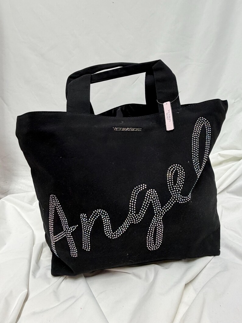 May include: Black tote bag with the word "Angel" spelled out in rhinestones. The bag has two handles and the brand name "Victoria's Secret" is visible. A pink tag is attached to one of the handles.