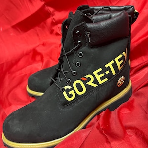 以下が含まれることがあります： 黄色い「GORE-TEX」の文字とトリムが付いた黒い革のブーツ。ブーツには黒い靴紐、金属製のアイレット、金のエンブレムが付いています。ブーツは赤い背景にあります。