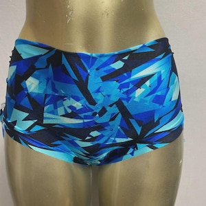 May include: Blue geometric print shorts. The shorts feature a high waist and a vibrant pattern of blue, black, and white shapes. The fabric appears to be a smooth, possibly athletic material. The shorts are displayed on a mannequin.