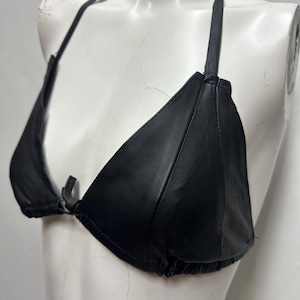 May include: Black leather bikini top with triangle cups and halter neck ties. The top is displayed on a white mannequin. The bikini top has a tie closure at the front and a gathered bottom edge.