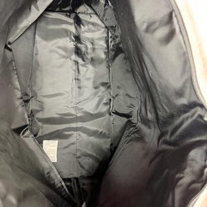 May include: Interior view of a large, open tote bag. The inside is lined with black, shiny fabric. The bag's exterior is a light beige color. The bag is empty and the zipper is open.