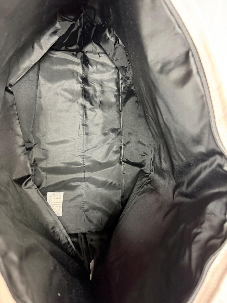 May include: Interior view of a large, open tote bag. The inside is lined with black, shiny fabric. The bag's exterior is a light beige color. The bag is empty and the zipper is open.