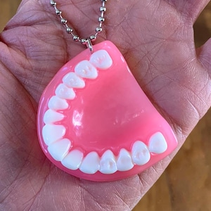 May include: A novelty necklace with a pink denture pendant and white teeth, suspended from a silver ball chain. The necklace is held in a hand.