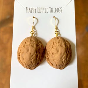 Puede incluir: Un par de pendientes novedosos con forma de nuez, con ganchos dorados. Los pendientes son de color marrón claro y se exhiben en una tarjeta blanca con el texto "HAPPY LITTLE THINGS".