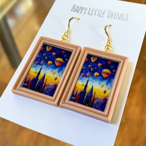 May include: A pair of earrings featuring miniature framed art with a hot air balloon scene. The frames are a light brown color, and the hooks are gold. The background of the art is a blue sky with colorful hot air balloons.