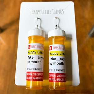 Könnte beinhalten: Ein Paar gelbe Pillenfläschchen-Ohrringe mit dem Text "Happy Little Things" und "Happy Little" auf den Etiketten. Die Etiketten zeigen auch "Take_table by mouth" und "Refills: Unlimited".