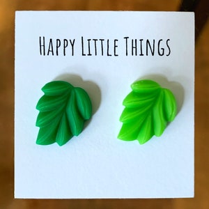 Pode incluir: Um par de brincos em forma de folha verde em um cartão branco com o texto "HAPPY LITTLE THINGS".