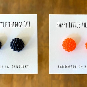May include: Two pairs of earrings on white cards. The cards have the text "Happy Little Things 101" and "Handmade in Kentucky". One pair of earrings is black and the other pair is orange.