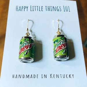 May include: A pair of handmade earrings featuring miniature Mountain Dew soda cans. The earrings have gold-colored hooks and are displayed on a white card with the text "HAPPY LITTLE THINGS 101" and "HANDMADE IN KENTUCKY".
