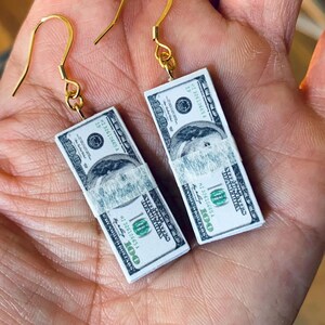 May include: A pair of gold earrings with stacks of $100 bills hanging from them. The bills have the text "HI 32610665 A" and "100" printed on them.
