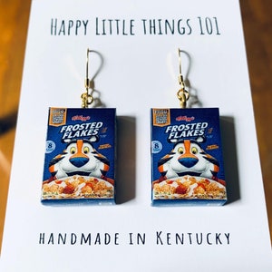 Puede incluir: Un par de pendientes con cajas de cereales en miniatura con la imagen de Tony el Tigre y el texto "Frosted Flakes". Los pendientes están hechos a mano en Kentucky.
