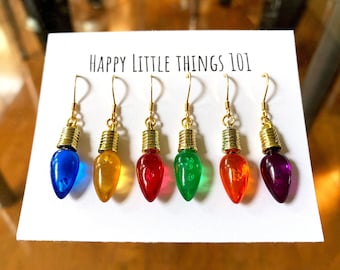 May include: A set of six colorful light bulb earrings with gold-colored metal accents. The earrings are arranged on a white card with the text "HAPPY LITTLE THINGS 101". Each bulb is a different color: blue, yellow, red, green, orange, and purple.
