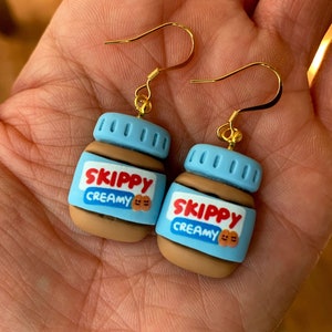 May include: A pair of gold dangle earrings featuring a peanut butter jar design. The jars are brown with a blue lid and the text "SKIPPY CREAMY" in red and white.
