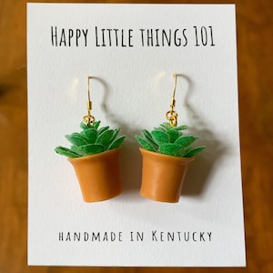 May include: A pair of gold dangle earrings featuring small potted succulent plants. The earrings are handmade in Kentucky and have the text "HAPPY LITTLE THINGS 101" on the card.
