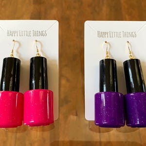 May include: Two pairs of dangle earrings shaped like nail polish bottles. One pair is pink with glitter and the other pair is purple with glitter. The earrings are on white cards with the text "Happy Little Things" printed on them.