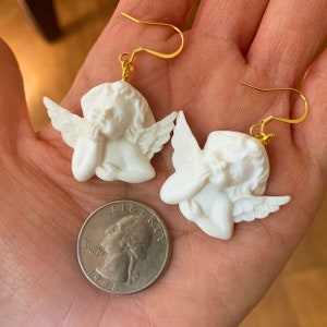 May include: A pair of gold angel earrings with white cherub figures. The cherubs are depicted with their hands covering their faces, and each has a pair of wings. The earrings are shown on a hand holding a US quarter.