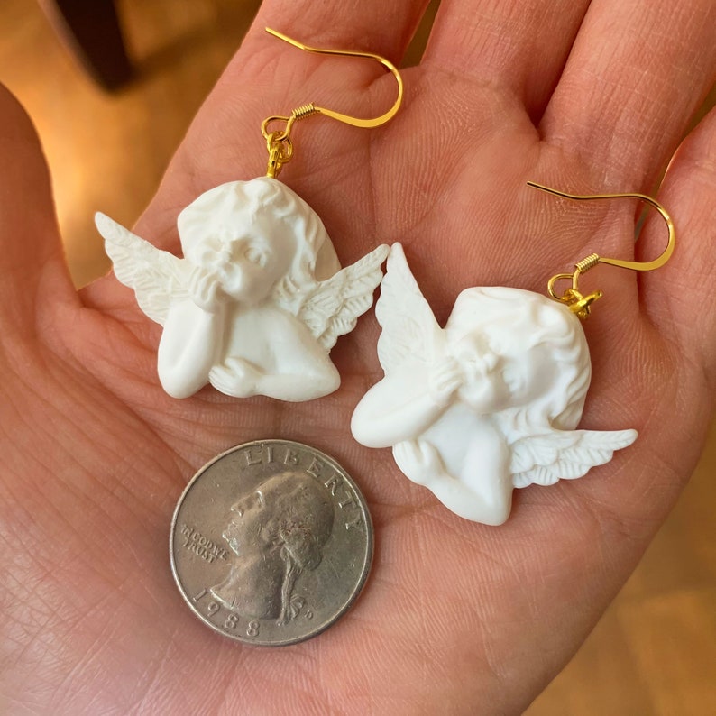 May include: A pair of gold angel earrings with white cherub figures. The cherubs are depicted with their hands covering their faces, and each has a pair of wings. The earrings are shown on a hand holding a US quarter.