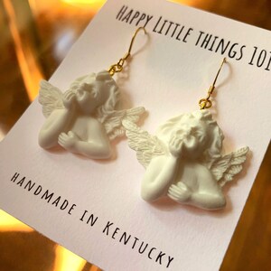 May include: White ceramic angel earrings with gold hooks. The angels are depicted with wings and have their hands clasped together. The earrings are handmade in Kentucky and are labeled "Happy Little Things 101".