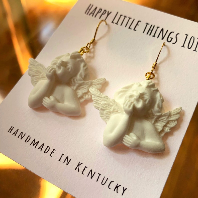 May include: White ceramic angel earrings with gold hooks. The angels are depicted with wings and have their hands clasped together. The earrings are handmade in Kentucky and are labeled "Happy Little Things 101".