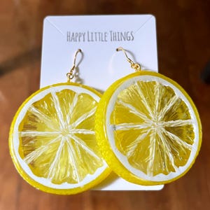 Può includere: Un paio di orecchini a forma di fetta di limone con segmenti gialli traslucidi, dettagli della buccia bianchi e ganci in metallo dorato. Gli orecchini sono fissati su un cartoncino bianco con la scritta Happy Little Things in alto.
