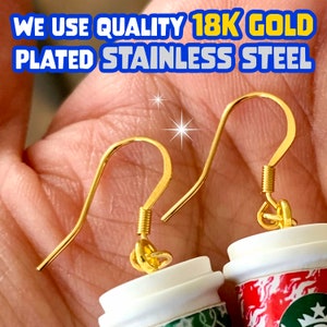 May include: A pair of gold-plated stainless steel earrings featuring miniature coffee cups. The cups are white with a green and red design. The earrings are in the shape of a hook.