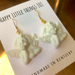 May include: White ceramic angel earrings with gold hooks. The angels are depicted with wings and have a thoughtful expression. The earrings are handmade in Kentucky. The text "HAPPY LITTLE THINGS 101" is visible on the card.