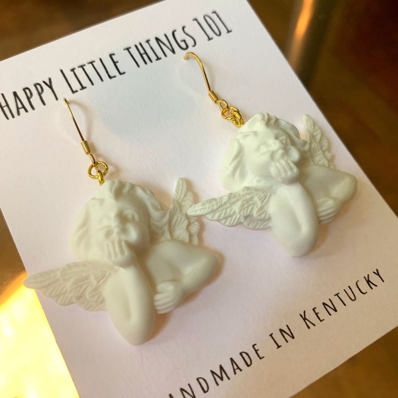 May include: White ceramic angel earrings with gold hooks. The angels are depicted with wings and have a thoughtful expression. The earrings are handmade in Kentucky. The text "HAPPY LITTLE THINGS 101" is visible on the card.