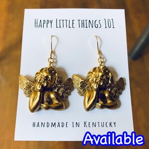 May include: Gold angel cherub dangle earrings with a textured finish. The earrings are handmade in Kentucky. The text "HAPPY LITTLE THINGS 101" and "HANDMADE IN KENTUCKY" are visible on the white card behind the earrings. The text "Available" is visible in blue at the bottom of the image.