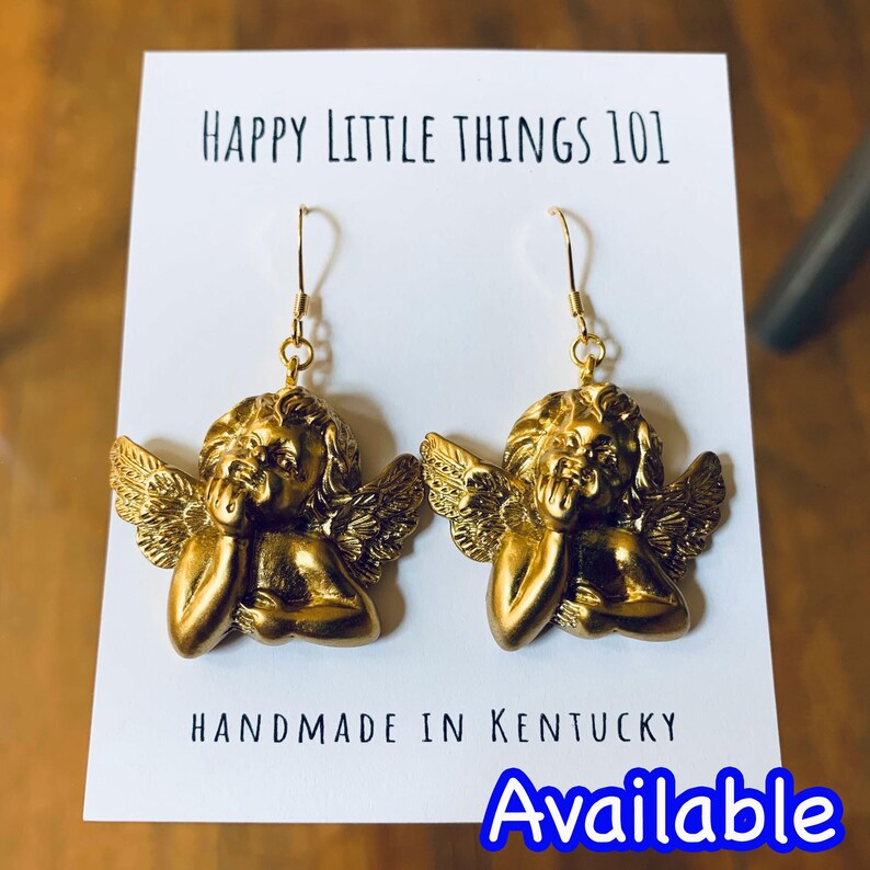 May include: Gold angel cherub dangle earrings with a textured finish. The earrings are handmade in Kentucky. The text "HAPPY LITTLE THINGS 101" and "HANDMADE IN KENTUCKY" are visible on the white card behind the earrings. The text "Available" is visible in blue at the bottom of the image.