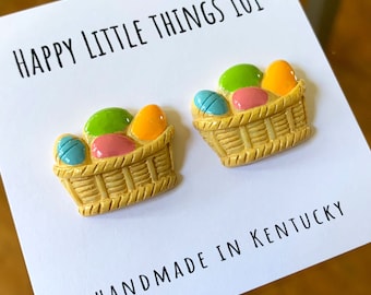 Könnte beinhalten: Ein Paar handgefertigte Keramikohrringe mit einem Korb voller bunter Ostereier. Die Ohrringe sind gelb mit braunen Korbdetails und tragen die Aufschrift "Happy Little Things 101" und "Handmade in Kentucky".