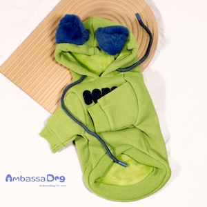 May include: Green hooded sweatshirt with blue fuzzy ear accents for a dog. The sweatshirt has a black embroidered logo on the front.
