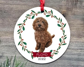 May include: A round white Christmas ornament with a brown dog inside a green wreath with red berries. The ornament has the text "FLOYD 2022" on the bottom.