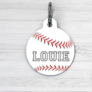 May include: White round pet ID tag with a baseball design. The tag has red stitching and the name "LOUIE" printed in black. The back of the tag has a phone number, address, and the text "Microchipped".