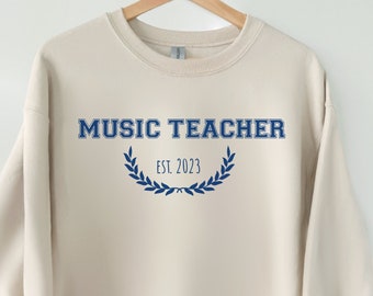 May include: A beige sweatshirt with the text "MUSIC TEACHER EST. 2023" in blue lettering with a blue laurel wreath.