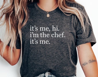 May include: A woman wearing a dark grey t-shirt with the text "it's me, hi. i'm the chef. it's me." in white lettering. She is wearing light blue jeans and white sneakers.
