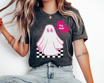 May include: Dark gray t-shirt featuring a pink ghost graphic with the text "Hey Boo!" in a pink heart-shaped speech bubble. The ghost is set against a black and white checkered pattern.