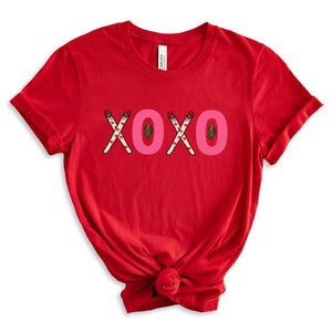 May include: Red short-sleeved t-shirt with the word "XOXO" printed in pink letters. The letters are made up of crossed candy canes with red hearts and green Christmas trees.
